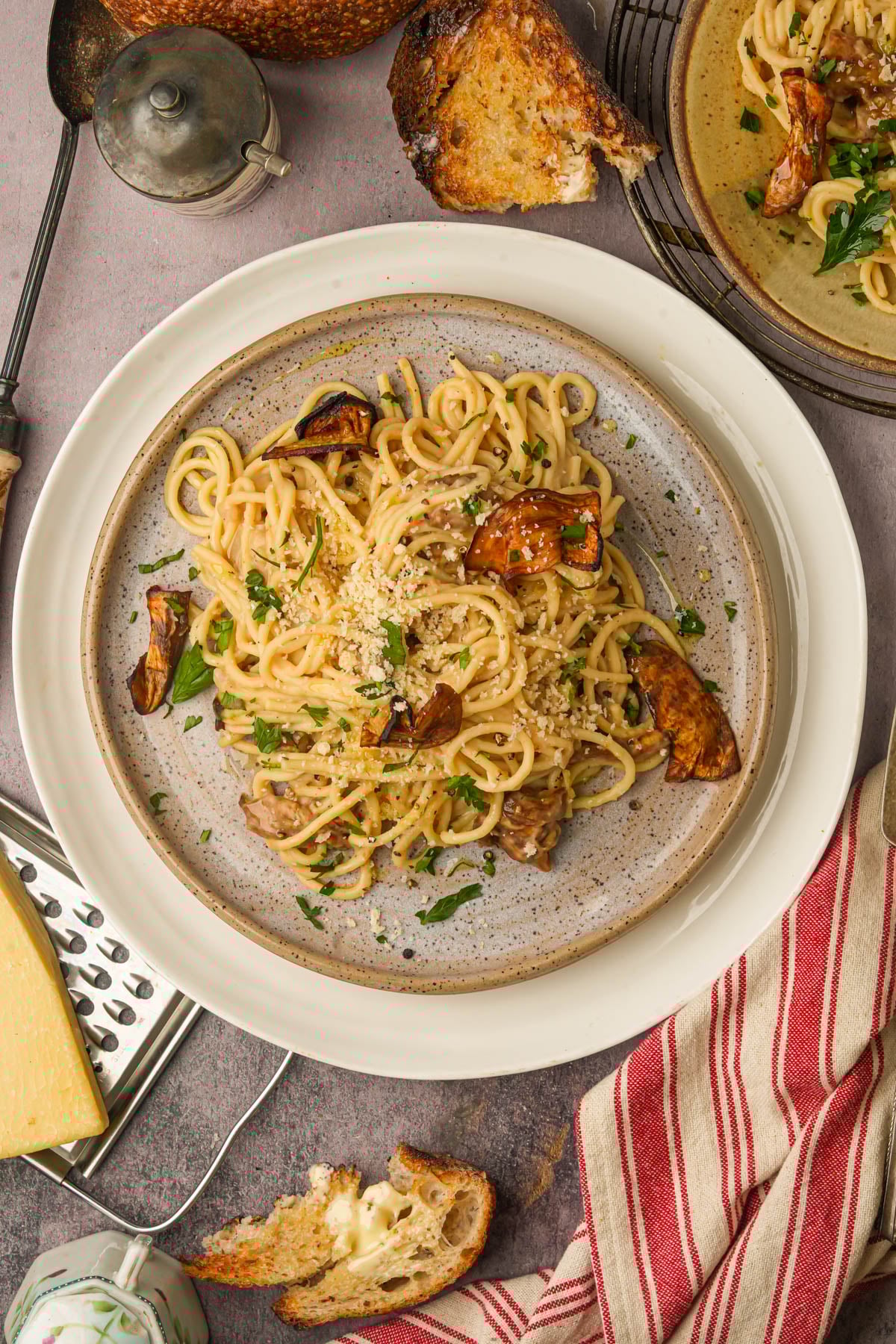 A plate of vegan carbonara pasta with eggplant, grated cheese, and parsley is on a table with toasted bread, a cheese grater, a wedge of cheese, and a red-striped napkin nearby. Another plate of pasta is partially visible.