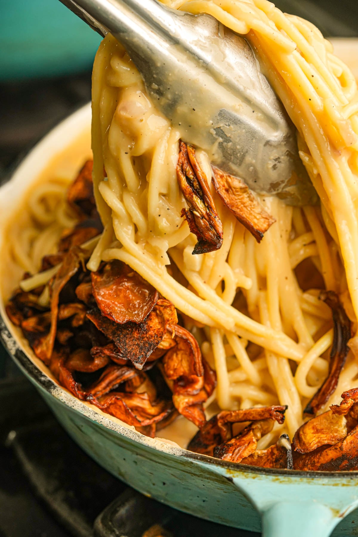Tongs lifting creamy vegan carbonara from a turquoise pot, with crispy roasted eggplant mixed in. The dish appears rich and savory.