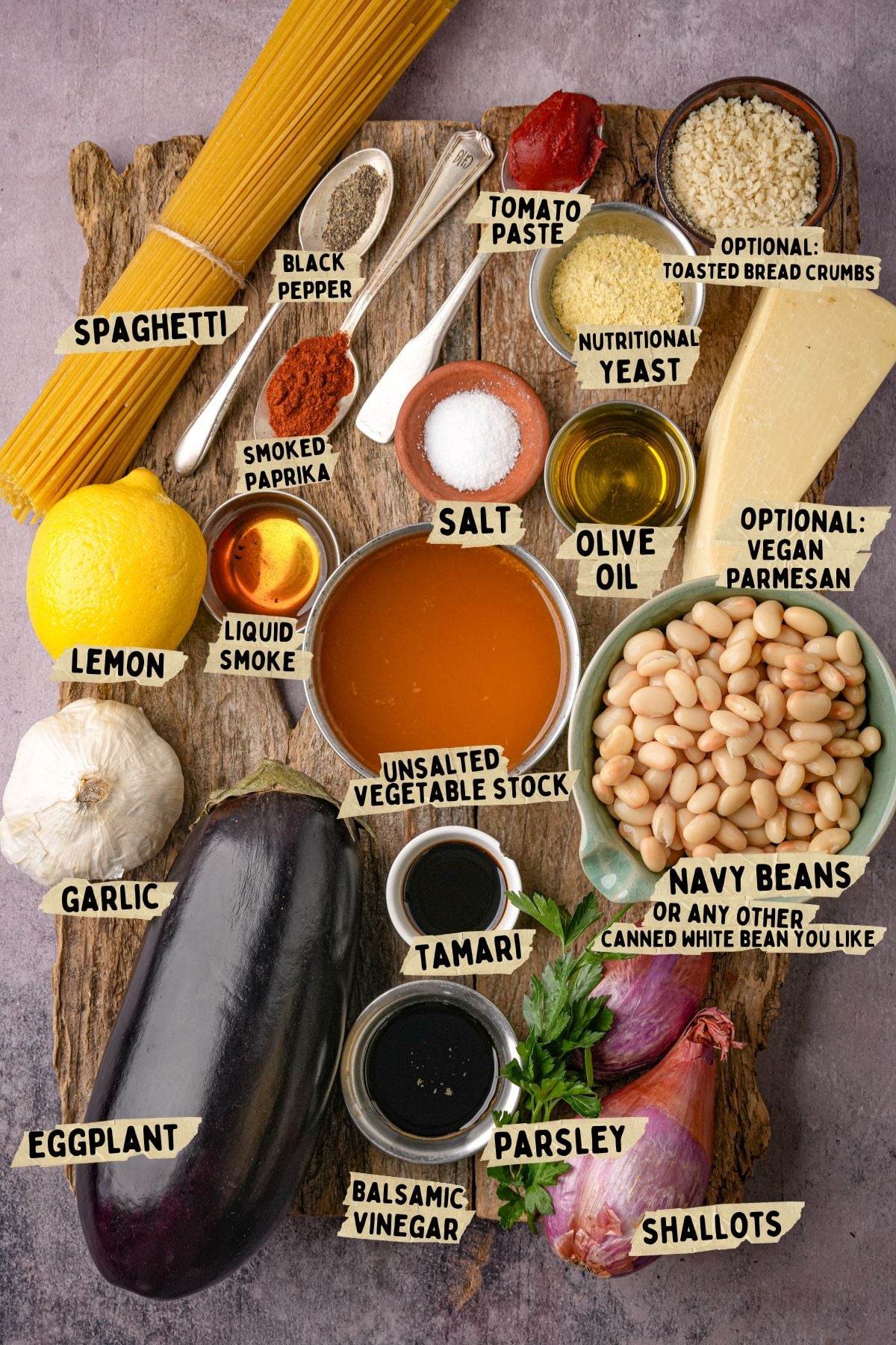 A top-down view of labeled ingredients for a vegan carbonara recipe on a wooden board, including spaghetti, eggplant, navy beans, garlic, shallots, lemon, olive oil, seasonings, tomato paste, vegan parmesan, breadcrumbs, and vegetable stock.