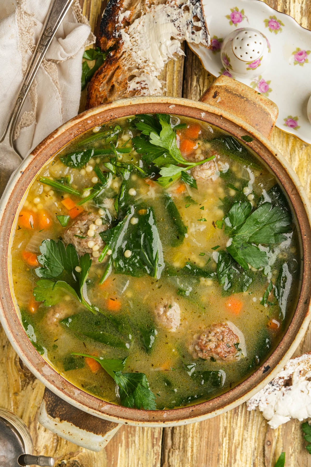A bowl of vegan Italian wedding soup with meatballs, carrots, spinach, orzo pasta, and parsley in a broth, served with slices of toasted bread on a rustic wooden table.
