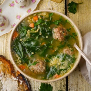 A bowl of vegan Italian wedding soup with meatballs, spinach, pasta, carrots, and parsley sits on a rustic wooden table next to buttered toast, a napkin, and floral salt and pepper shakers.
