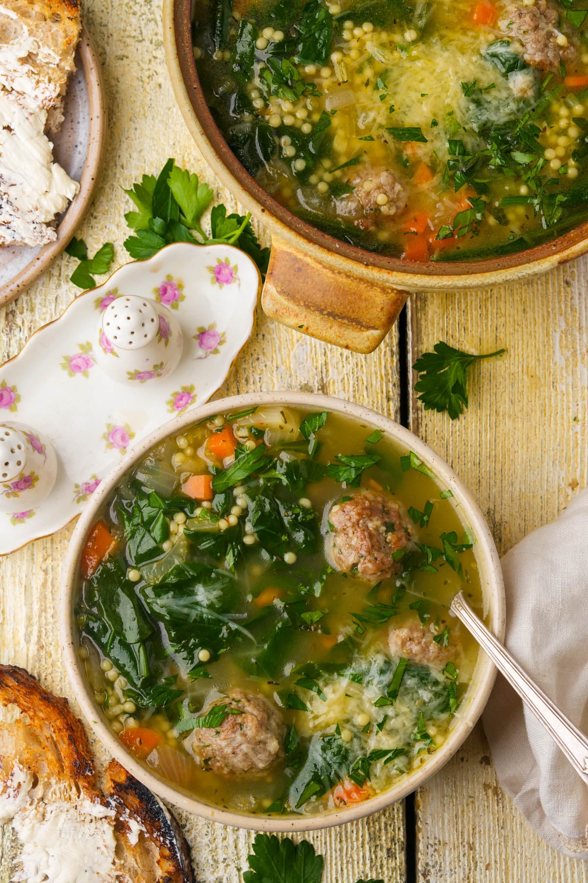 A bowl of vegan Italian wedding soup with meatballs, greens, carrots, pasta, and grated cheese sits on a rustic wooden table with toast, a partial soup pot, salt and pepper shakers, and a beige napkin nearby.