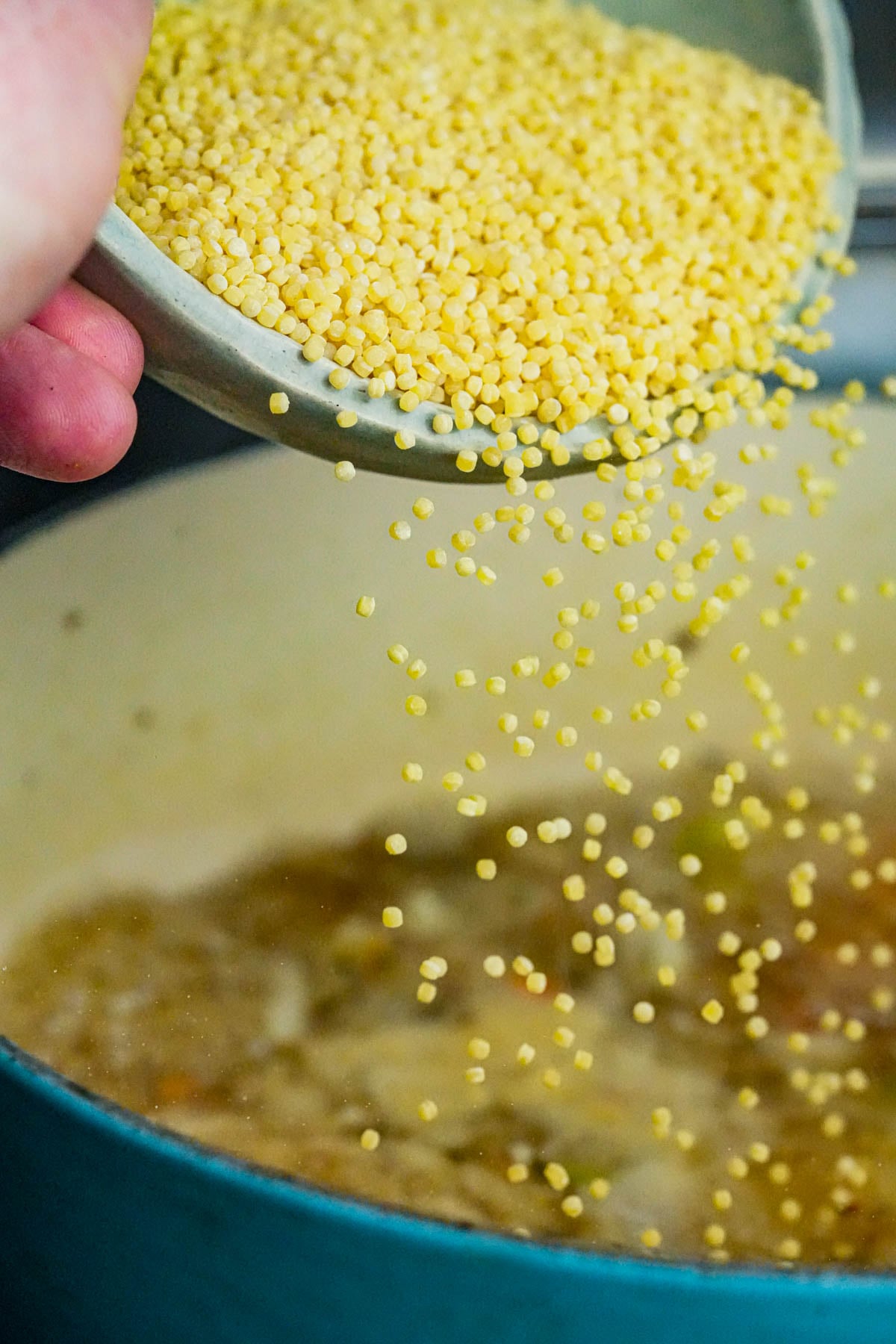 A hand pours acini de pepe from a bowl into a cooking pot filled with simmering vegan Italian wedding soup.