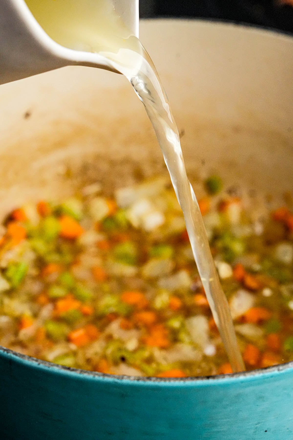 Broth being poured from a white pitcher into a pot with chopped vegetables, including carrots, celery, and onions, in a blue Dutch oven.