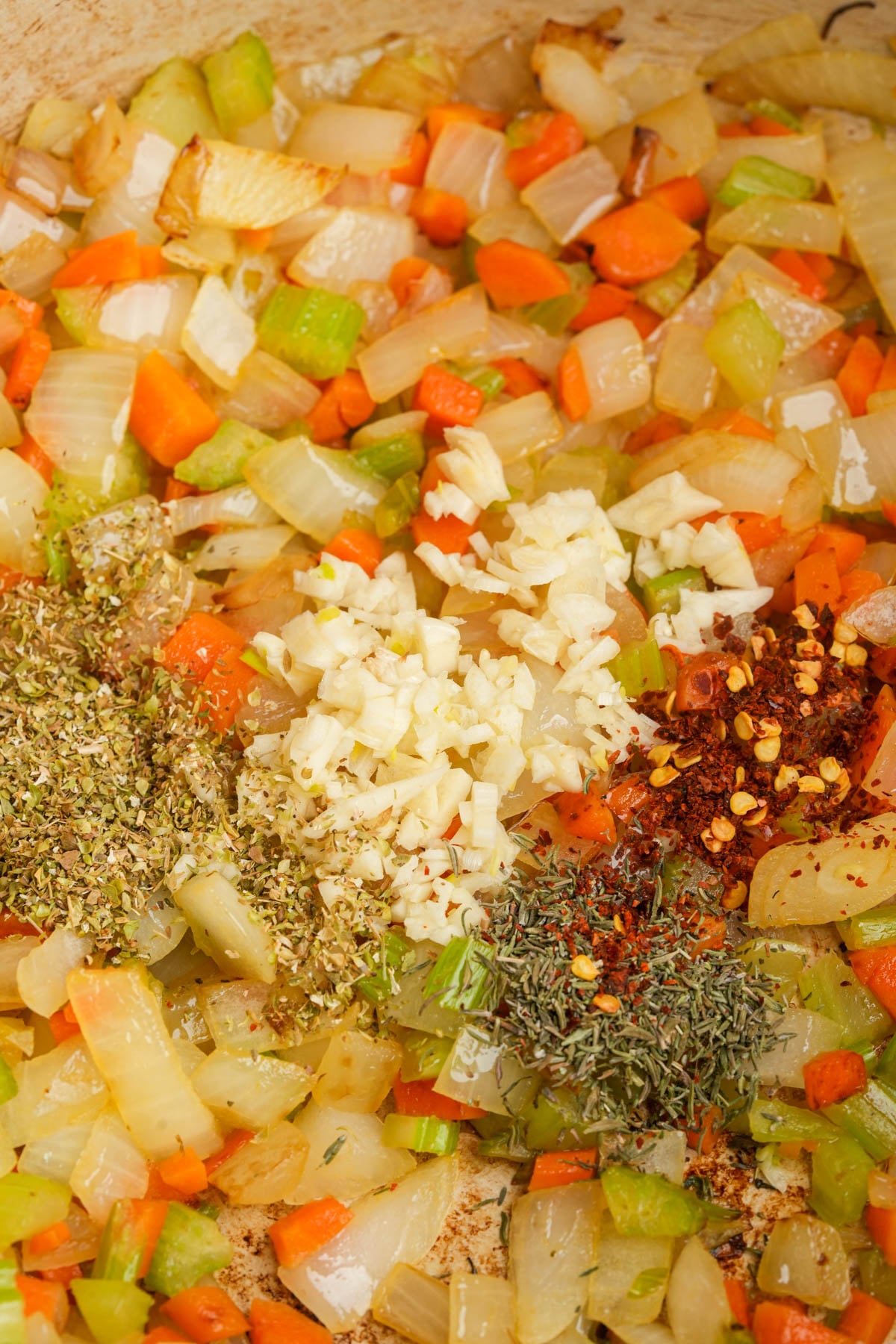Diced onions, carrots, and celery are sautéing in a pan. Piles of minced garlic, dried oregano, dried thyme, and crushed red pepper flakes are added on top, ready to be mixed in.