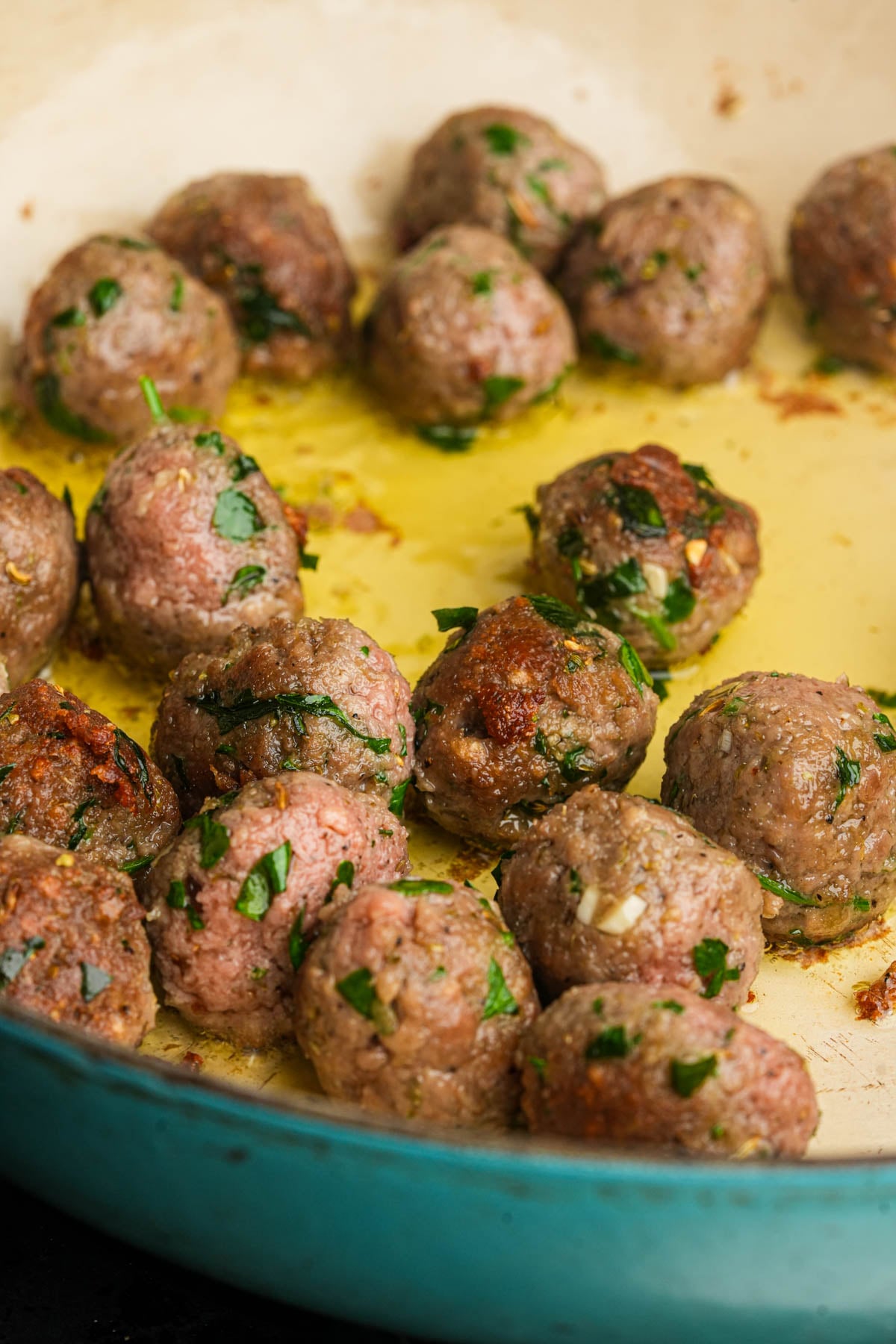 Meatballs with chopped herbs are being cooked in a shallow layer of oil in a blue skillet. Some meatballs are browned while others are still pink and in the process of cooking.
