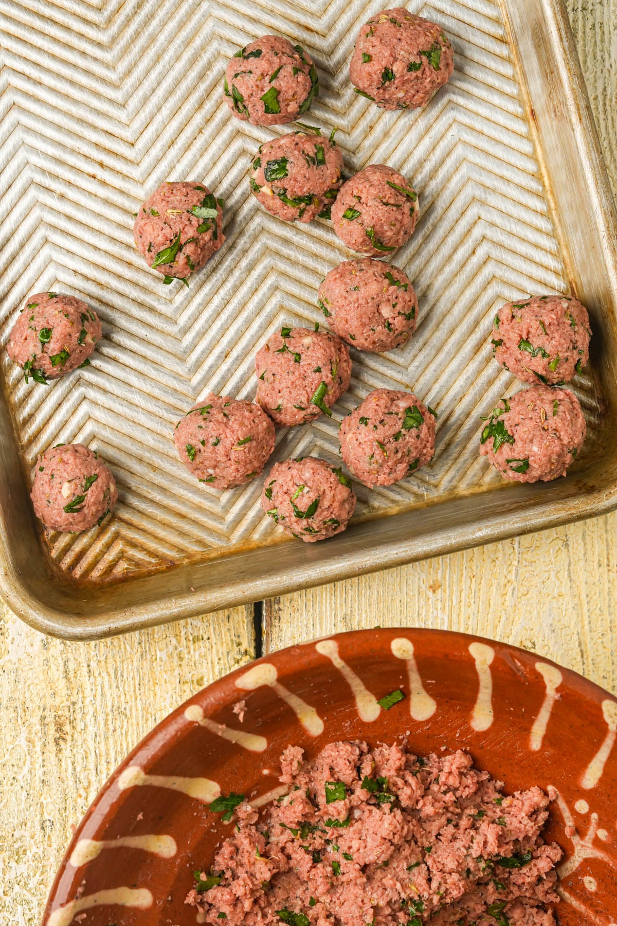 Raw meatballs with chopped herbs are arranged on a metal baking sheet. Below, a brown bowl contains the remaining meat mixture. The scene is set on a light wooden surface.