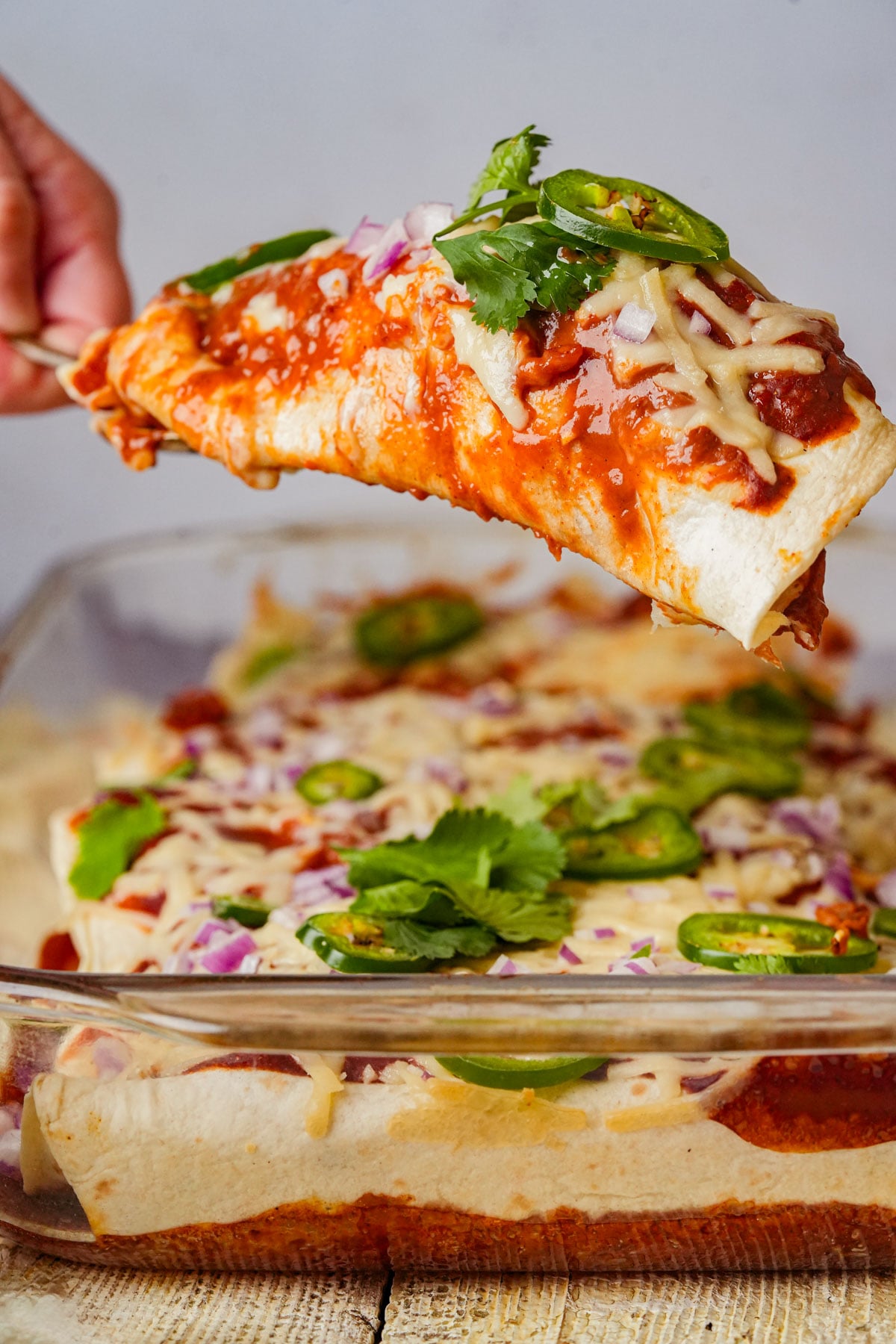 A close-up of a hand lifting a cheesy, saucy jackfruit enchilada from a glass baking dish, topped with cilantro, sliced jalapeños, and chopped red onions. Other enchiladas remain in the dish in the background.