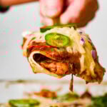 A close-up of a cheesy jackfruit enchilada filled with shredded jackfruit, topped with sliced jalapeños, red onions, and melted cheese, being lifted from a pan by a hand.