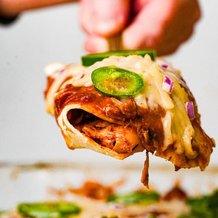 A close-up of a cheesy jackfruit enchilada filled with shredded jackfruit, topped with sliced jalapeños, red onions, and melted cheese, being lifted from a pan by a hand.