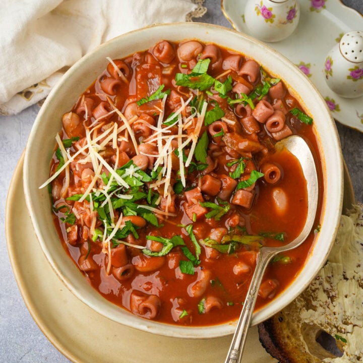 A bowl of tomato-based pasta soup with small pasta shapes, garnished with chopped herbs and shredded cheese, sits on a plate with a slice of bread and a silver spoon. A white napkin and teapot are in the background.