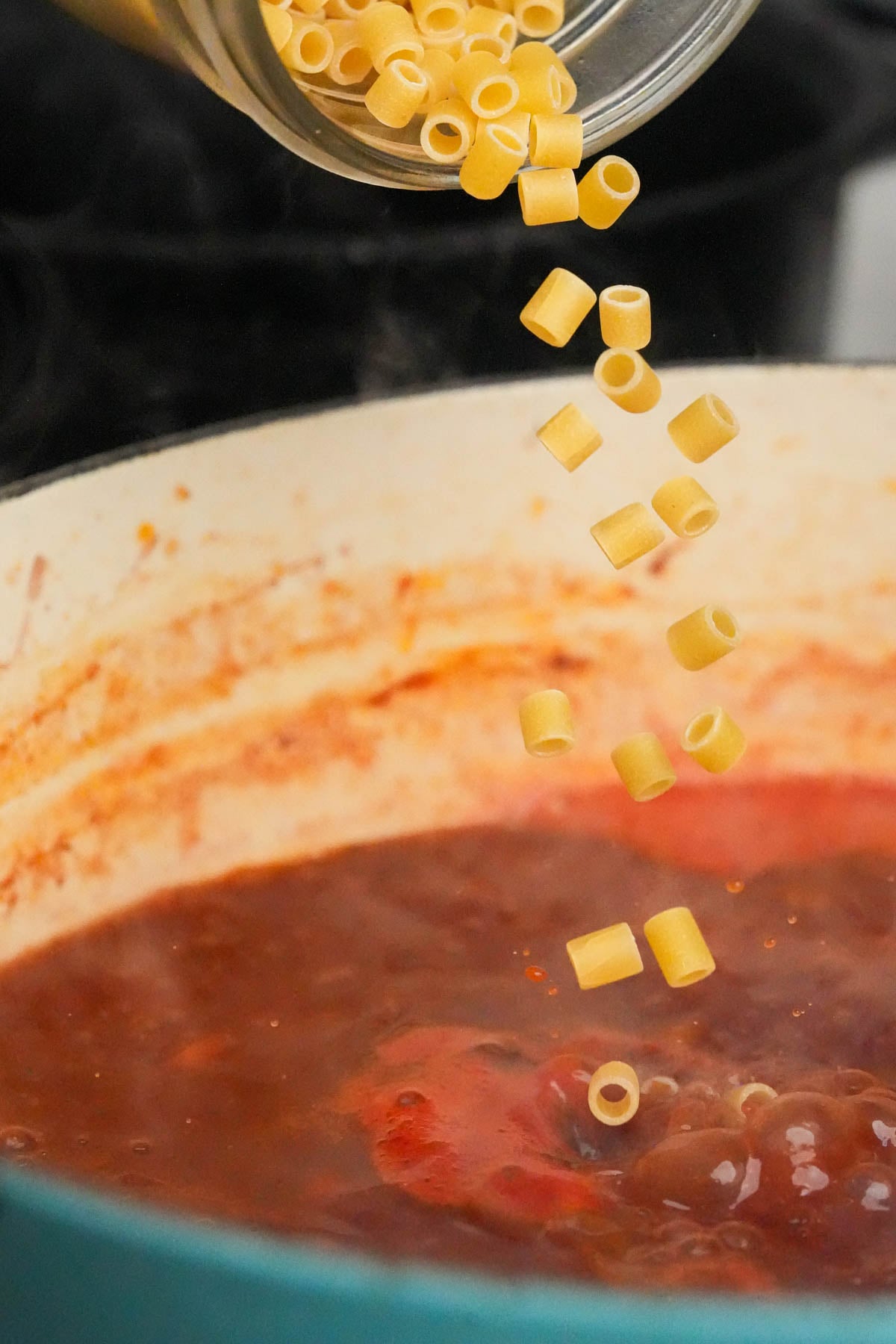 Uncooked ditalini pasta is being poured from a jar into a pot of simmering vegan pasta fagioli.
