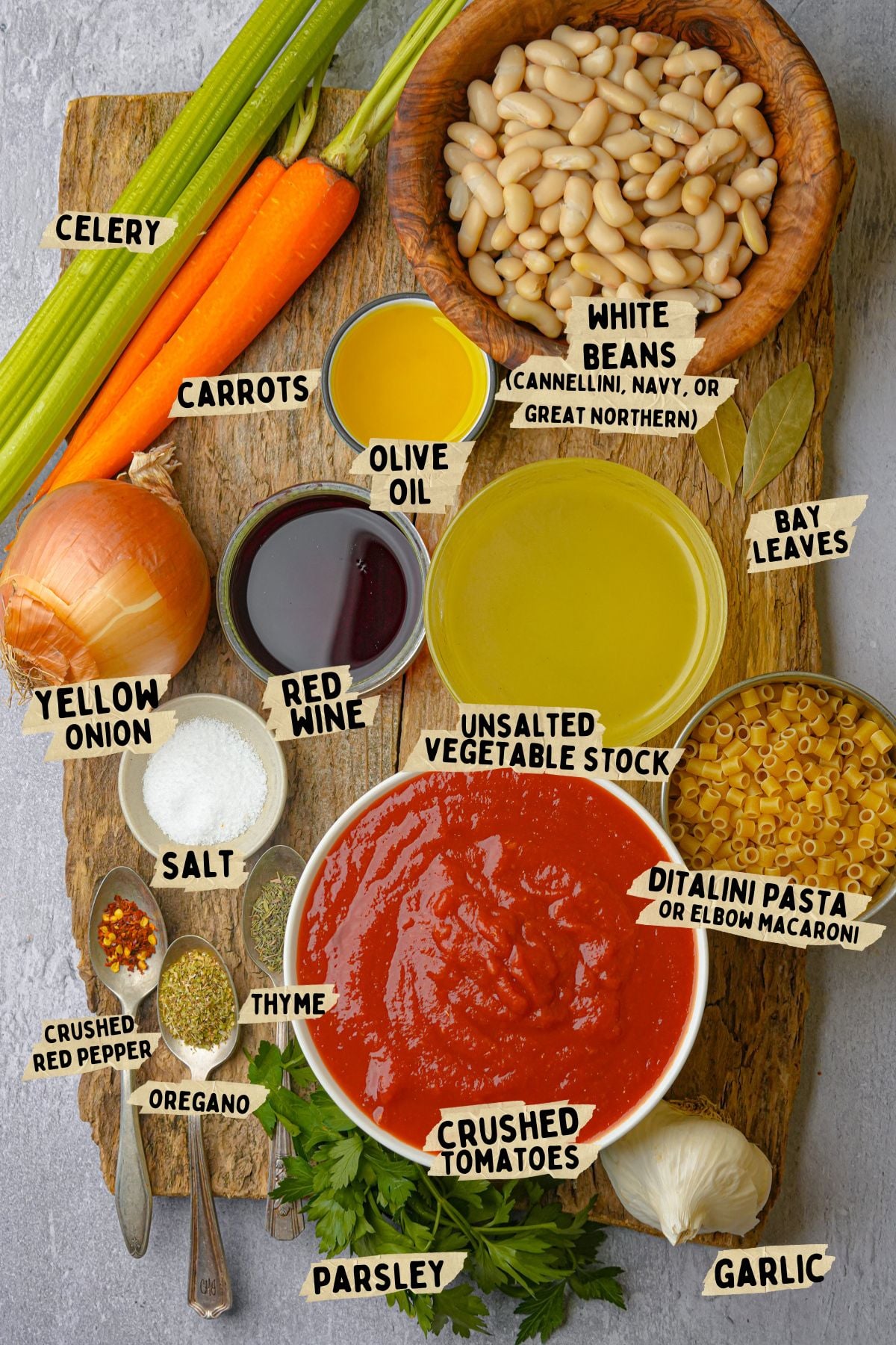 A flat lay of ingredients for soup: celery, carrots, white beans, olive oil, red wine, unsalted vegetable stock, bay leaves, ditalini pasta, garlic, parsley, crushed tomatoes, oregano, thyme, salt, and a yellow onion.