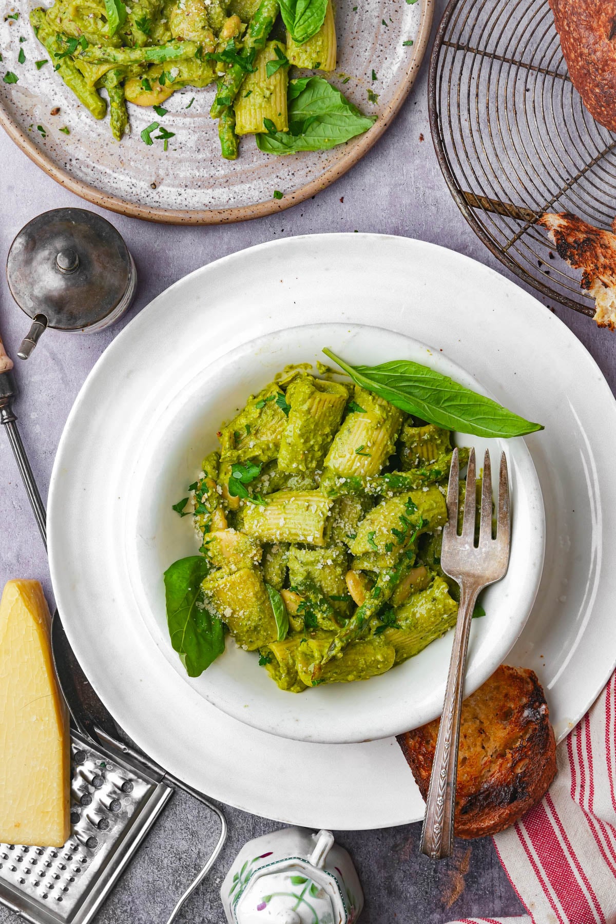 A bowl of vegan pesto rigatoni pasta coated in green pesto sauce, garnished with fresh basil and grated cheese. A fork rests on the side, with bread, cheese, a grater, and a plate of more pasta nearby on a light surface.