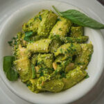 A white bowl filled with vegan pesto pasta coated in a creamy green pesto sauce, garnished with grated cheese and fresh basil leaves.