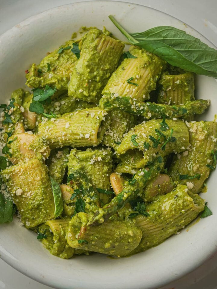 A white bowl filled with vegan pesto pasta coated in a creamy green pesto sauce, garnished with grated cheese and fresh basil leaves.