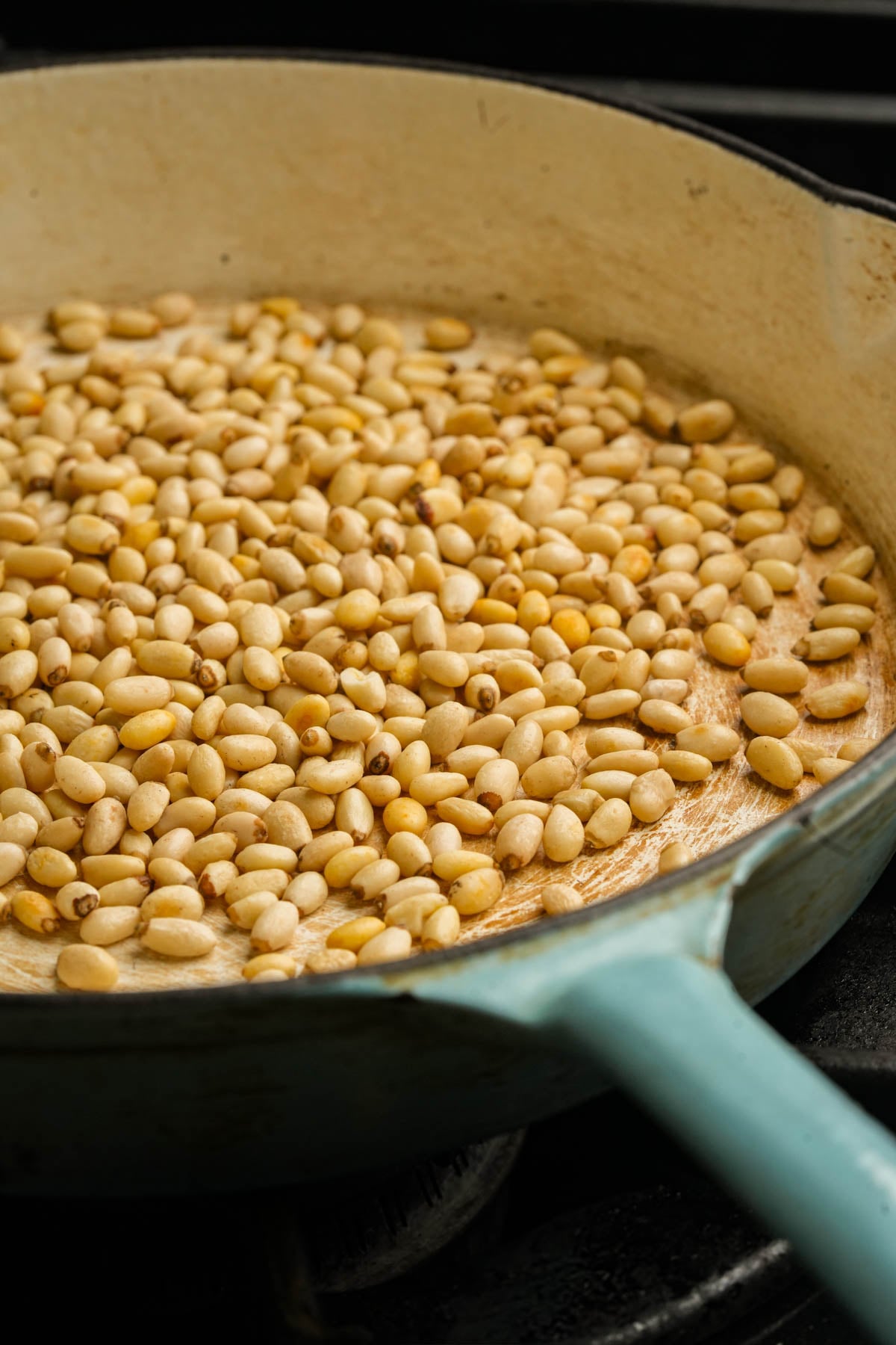 A close-up of pine nuts being toasted in a light-colored skillet with a blue handle on a stovetop.