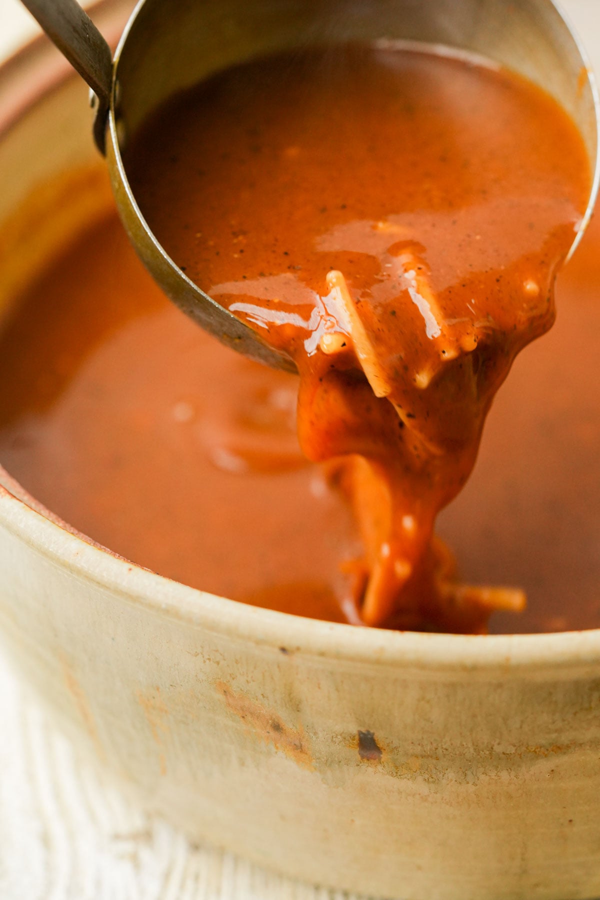 A ladle pours thick, rich sopa de fideo with visible spices and a few noodle pieces into a beige ceramic bowl.