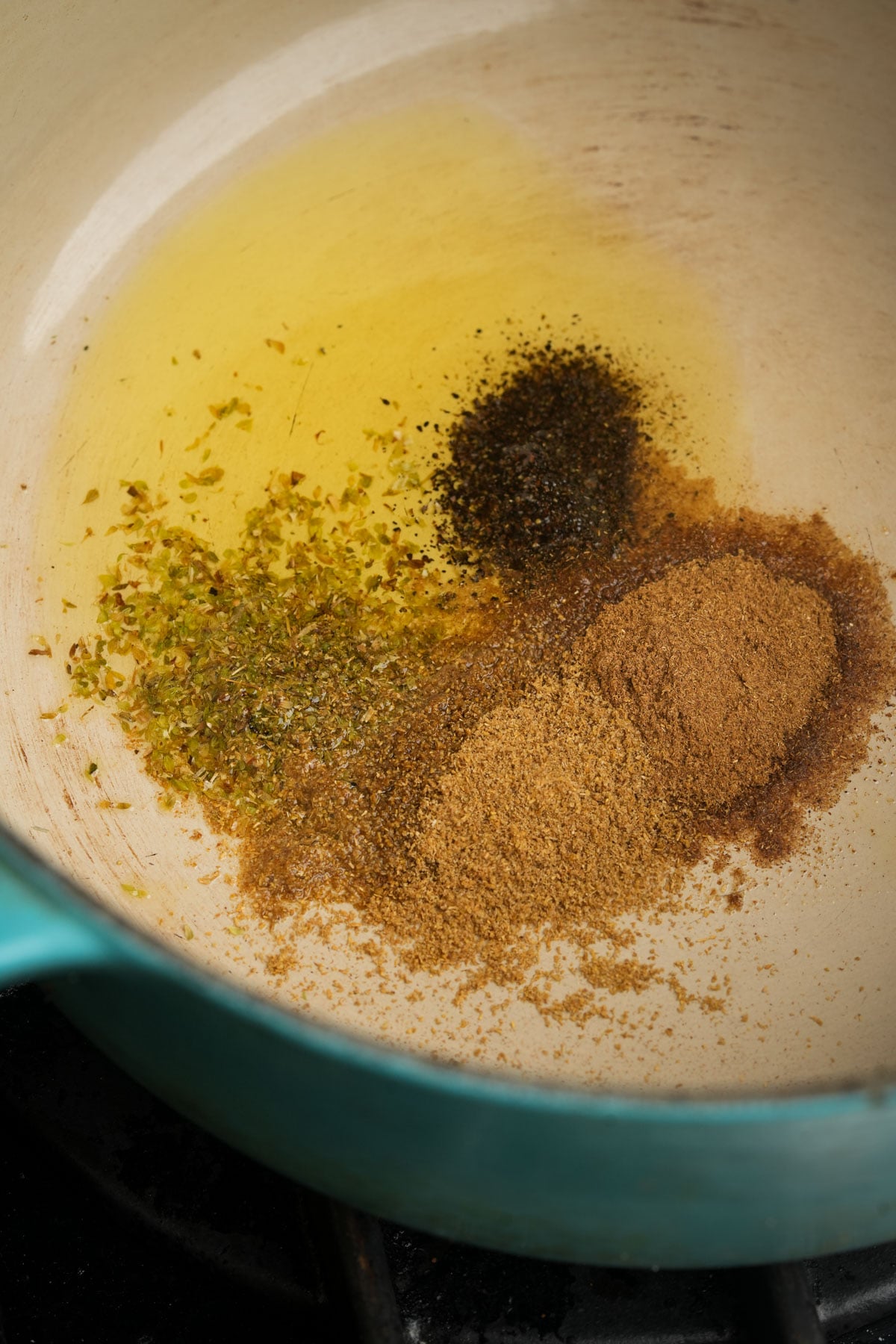 A turquoise pot with oil and four piles of spices, including ground coriander, cumin, black pepper, and dried oregano, being heated on a stovetop.