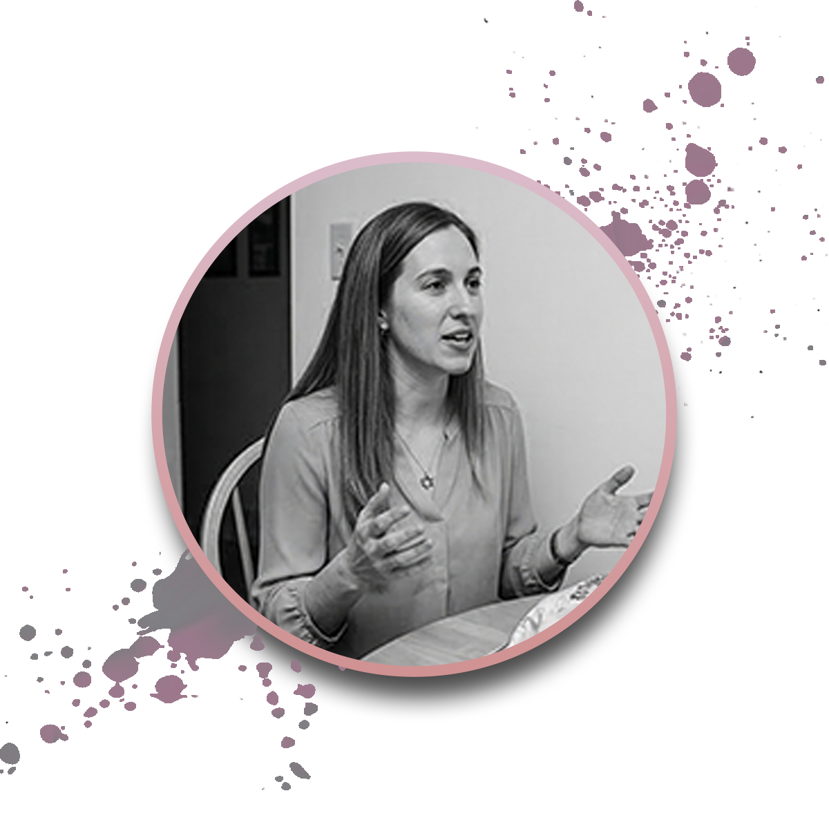 A woman with long hair gestures with her hands while sitting at a table. The photo is black and white with a pink circular border and paint splatter effects in the background.