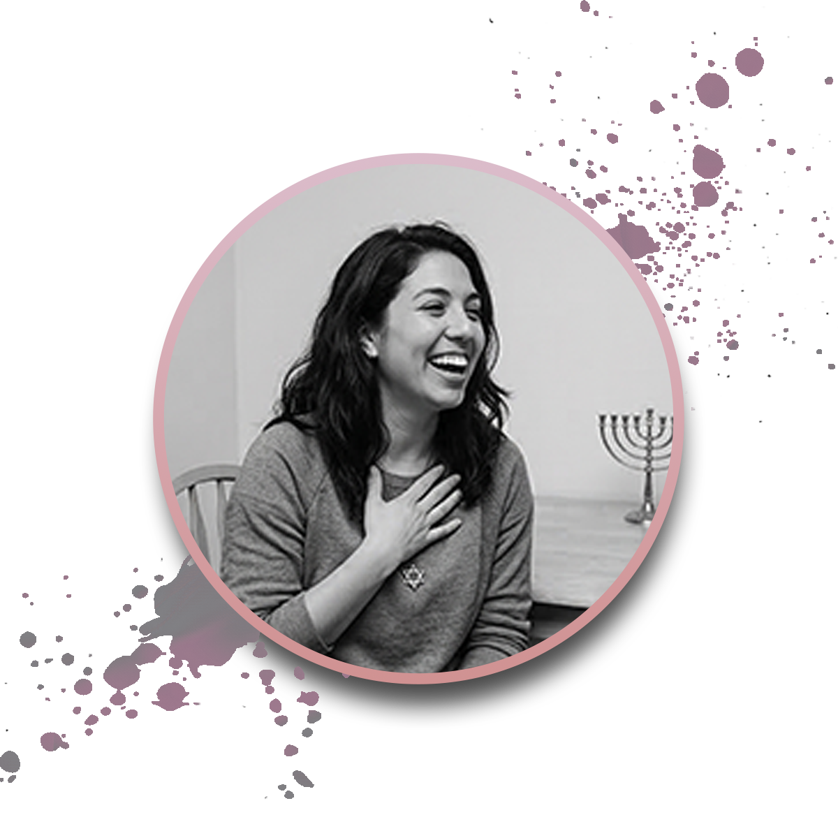 A woman with dark hair, smiling and holding her hand to her chest, sits at a table with a menorah in the background. The image is in black and white with a pink border and decorative splatters.