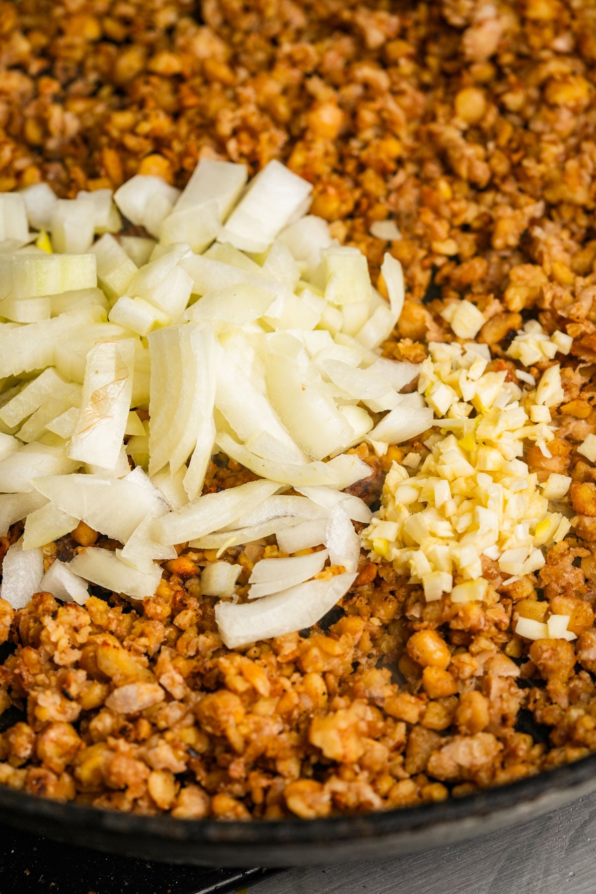 Chopped onions and minced garlic are added to a skillet filled with seasoned, browned ground chickpeas and walnut crumbles.