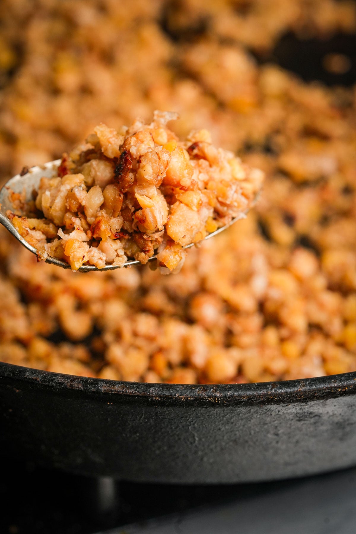 A close-up of a spoon holding a serving of browned, crumbled chickpea and walnut mixture over a skillet.