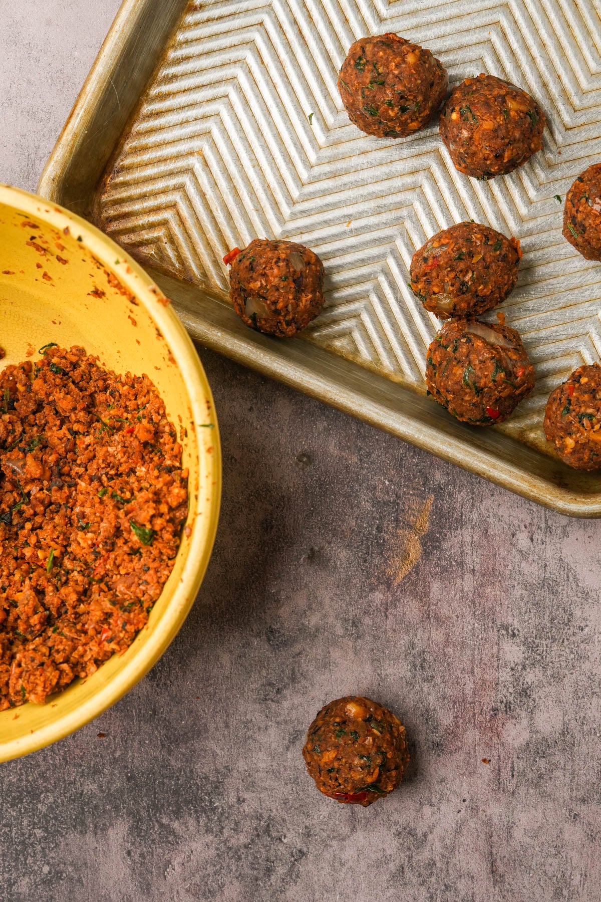 A yellow bowl of chickpea meatball mixture sits beside a baking tray with several formed falafel balls on a textured surface. One uncooked falafel ball rests on the surface near the bowl.