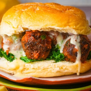 A close-up of a chickpea meatball filled with meatballs covered in tomato sauce, melted cheese, and fresh green leafy vegetables on a soft, golden bun.