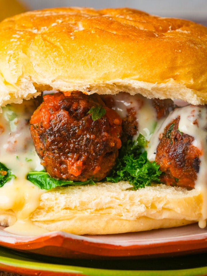 A close-up of a chickpea meatball filled with meatballs covered in tomato sauce, melted cheese, and fresh green leafy vegetables on a soft, golden bun.
