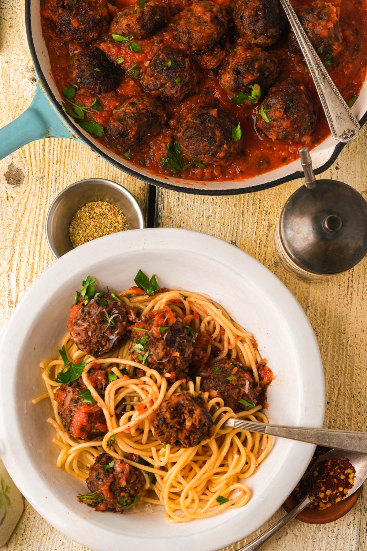 A bowl of spaghetti topped with chickpea meatballs and tomato sauce sits on a table next to a pan of more meatballs in sauce, with spices and a pepper grinder nearby.