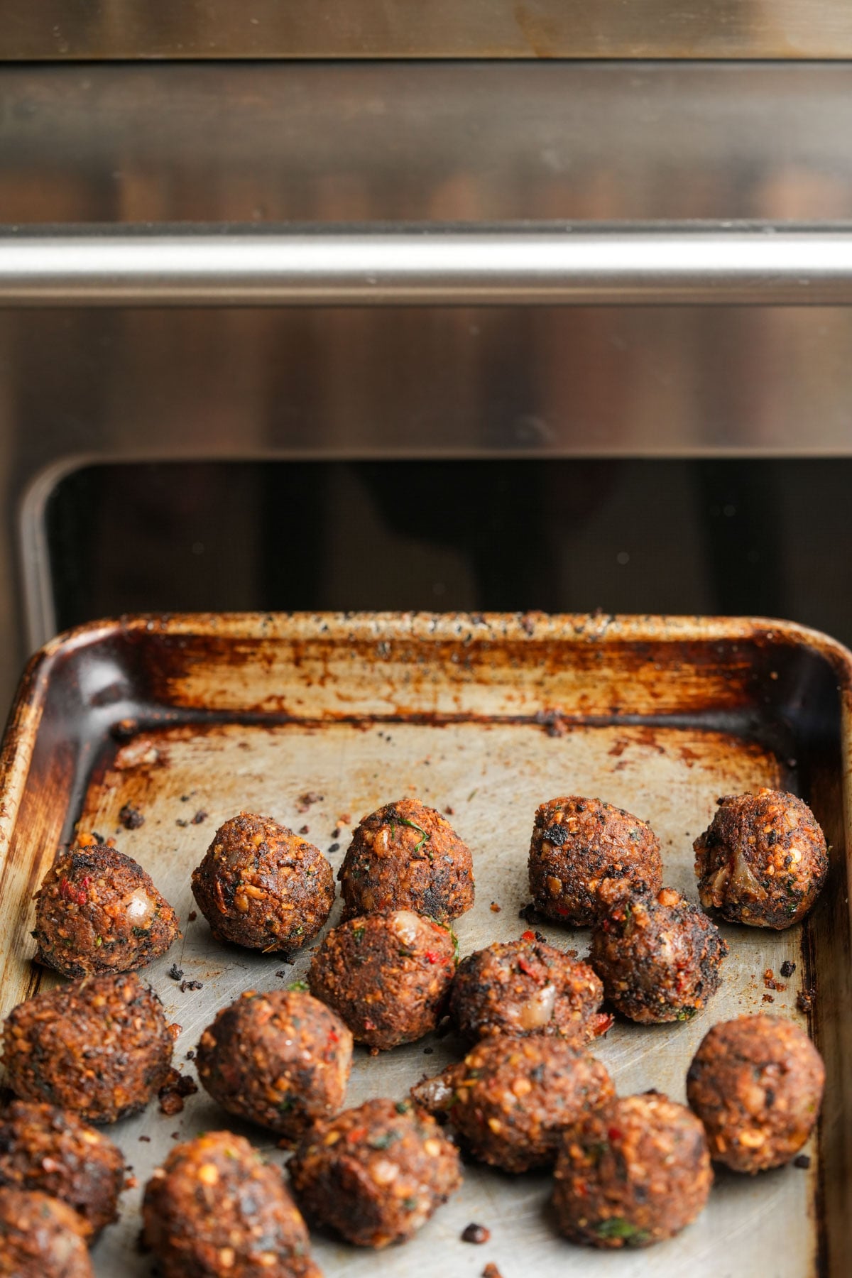 A metal baking sheet holds several browned, baked chickpea meatballs, some with visible herbs and bits of vegetables, set in front of a stainless steel oven.