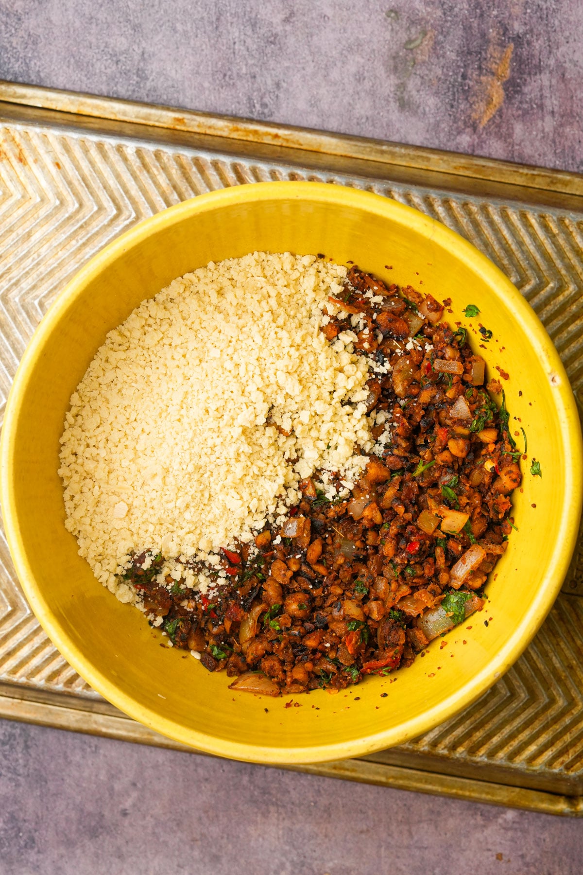 A yellow bowl contains dry, crumbled chickpea meatball mix on one side and breadcrumbs on the other, sitting on a metal tray with a textured pattern.