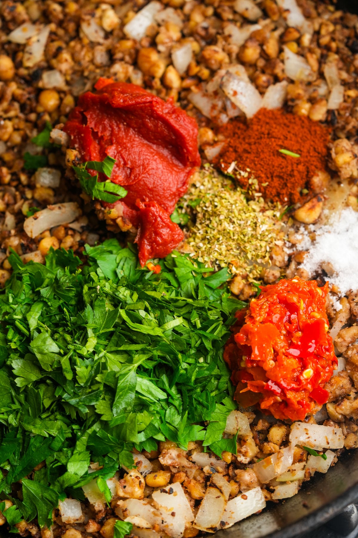 Chopped onions and chickpeas in a pan, topped with tomato paste, chopped parsley, red chili paste, dried herbs, red spices, and salt, about to be mixed together.