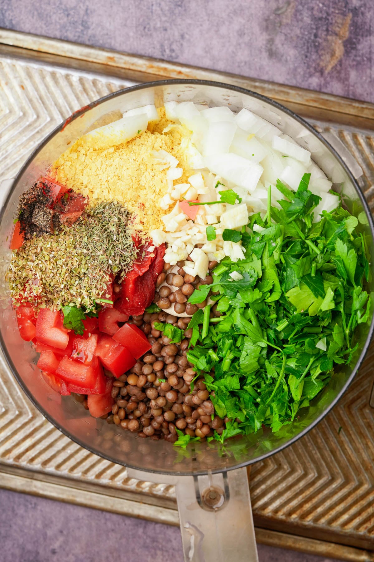 A food processor bowl filled with chopped onion, parsley, diced tomatoes, cooked lentils, nutritional yeast, tomato paste, garlic, dried herbs, and black pepper, all arranged in separate sections.