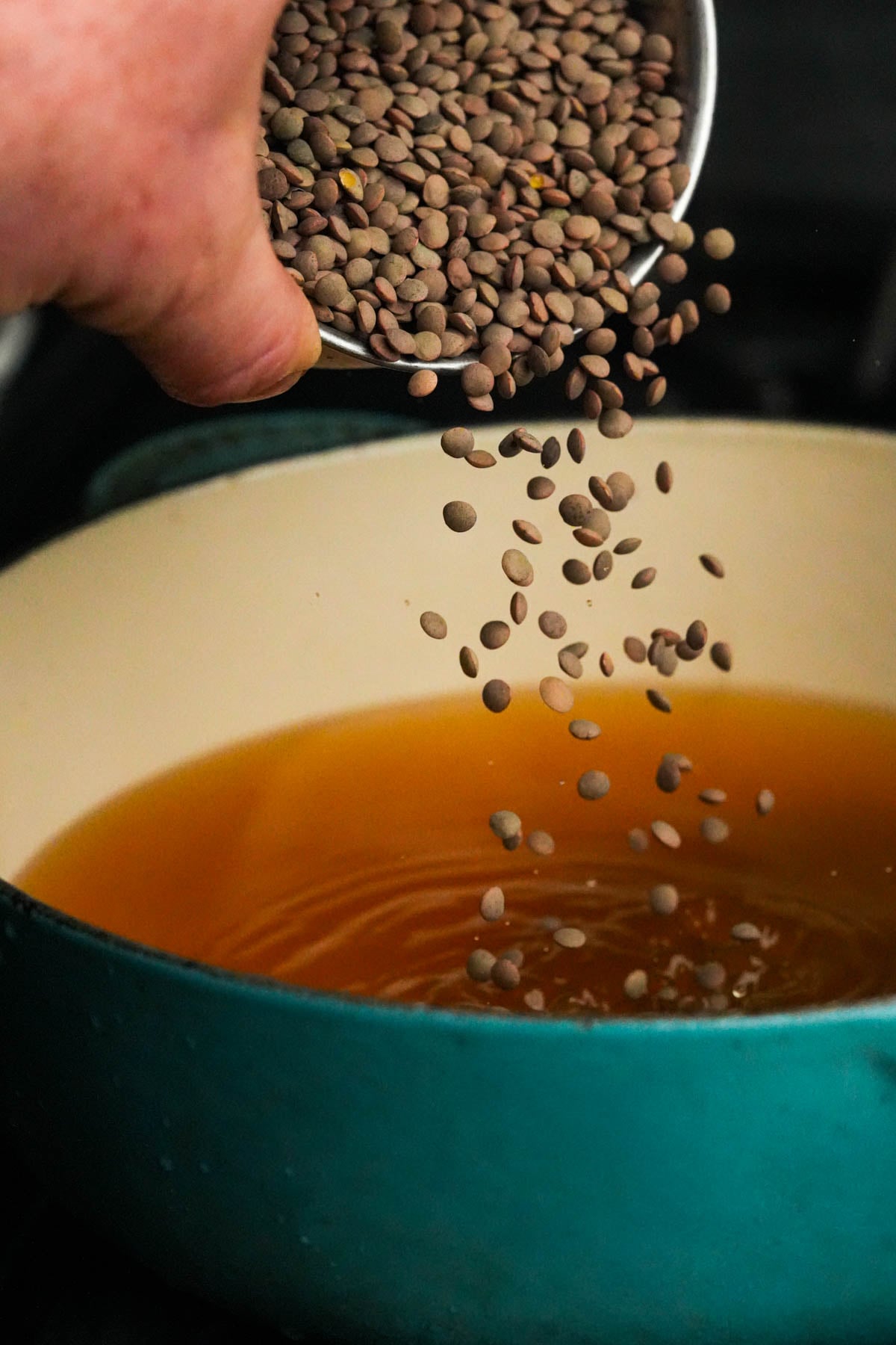 A hand pours dry lentils from a metal container into a teal pot filled with broth.