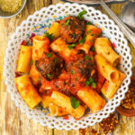 A decorative white plate filled with rigatoni pasta and lentil meatballs in tomato sauce, garnished with fresh parsley, set on a rustic wooden table with scattered red pepper flakes nearby.