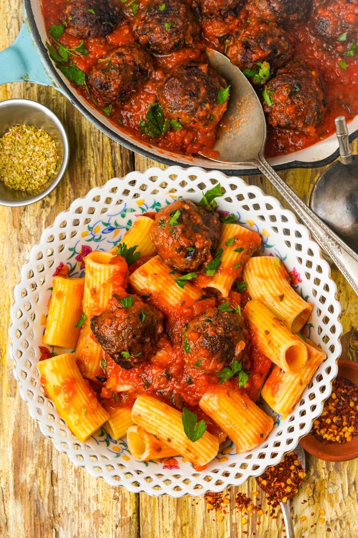 A bowl of rigatoni pasta topped with lentil meatballs and marinara sauce, garnished with fresh parsley. A skillet with more meatballs in sauce, a spoon, and small bowls of spices are nearby on a rustic wooden table.