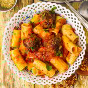 A decorative white plate filled with rigatoni pasta and lentil meatballs in tomato sauce, garnished with fresh parsley, set on a rustic wooden table with scattered red pepper flakes nearby.