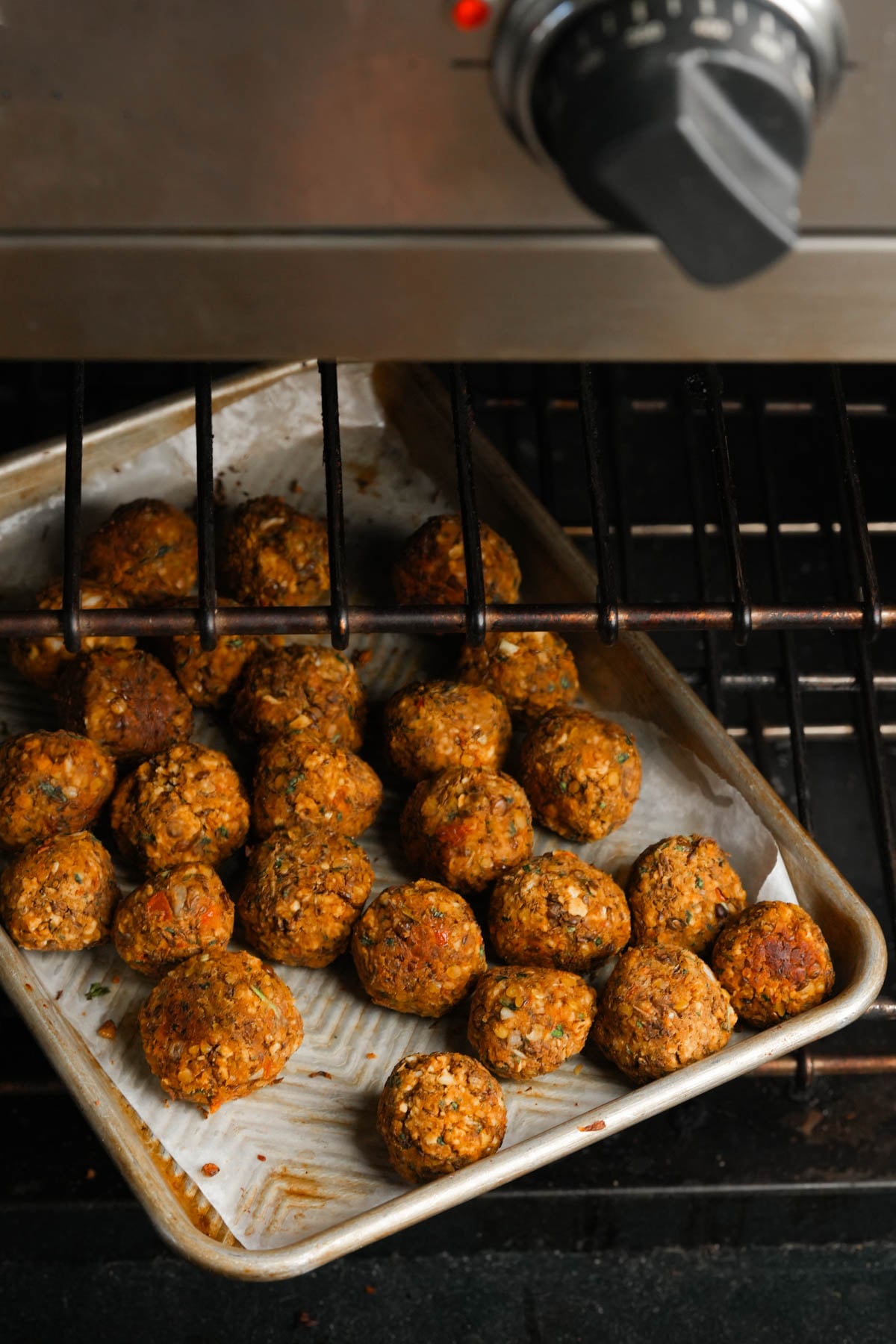 A baking tray lined with parchment paper holds several cooked lentil meatballs inside an oven, with the oven rack partially pulled out and the temperature dial visible at the top of the image.
