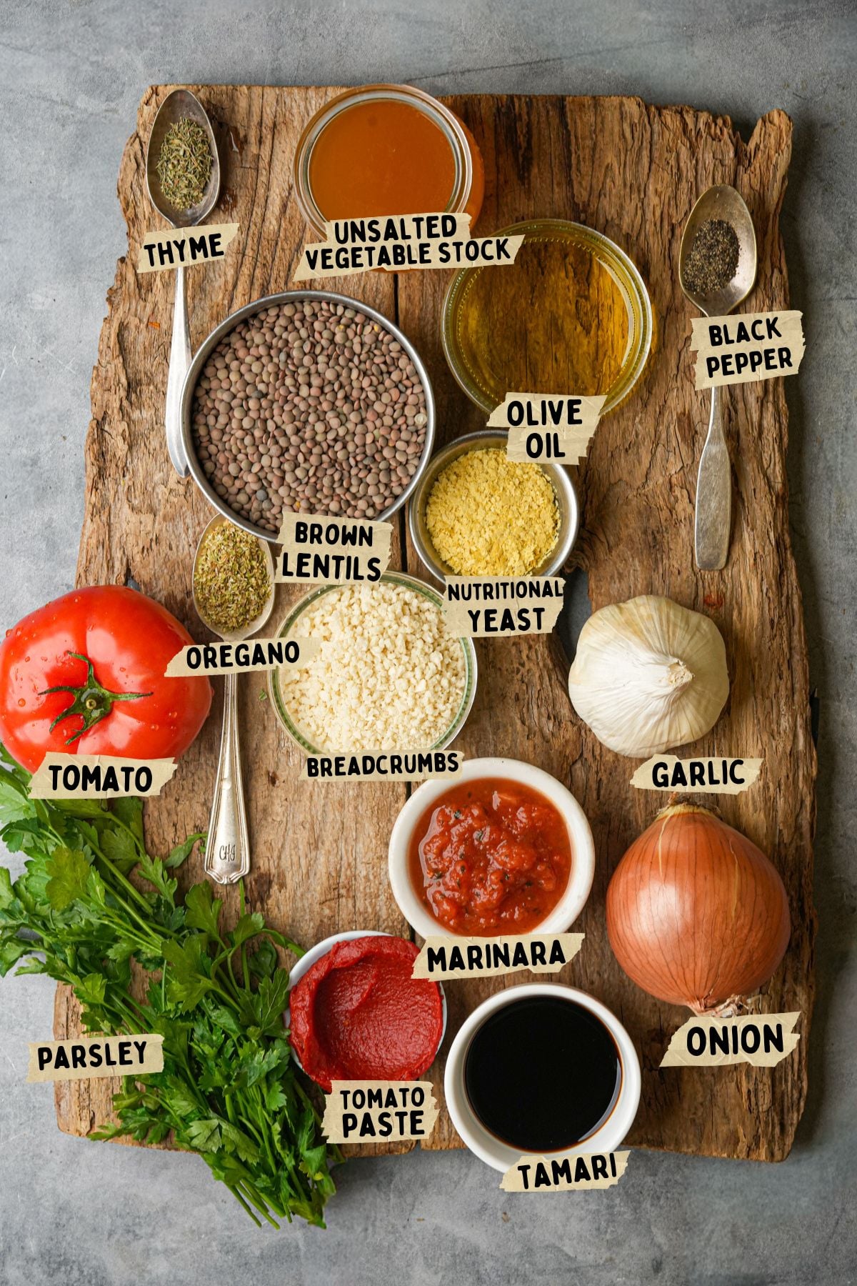 A wooden board displays labeled ingredients for a lentil meatballs recipe: thyme, unsalted vegetable stock, black pepper, olive oil, brown lentils, nutritional yeast, oregano, breadcrumbs, garlic, onion, parsley, tomato, marinara, tomato paste, and tamari.