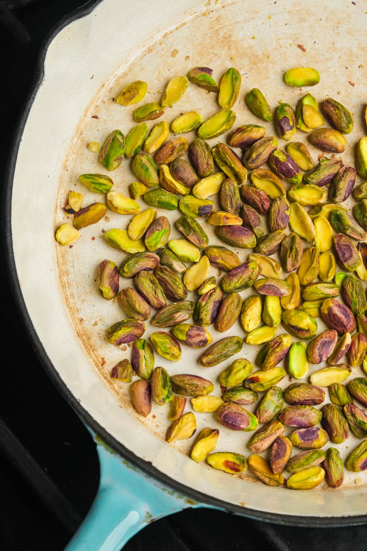 Shelled pistachios being roasted in a large, white enameled skillet with a light blue edge, sitting on a black stovetop.