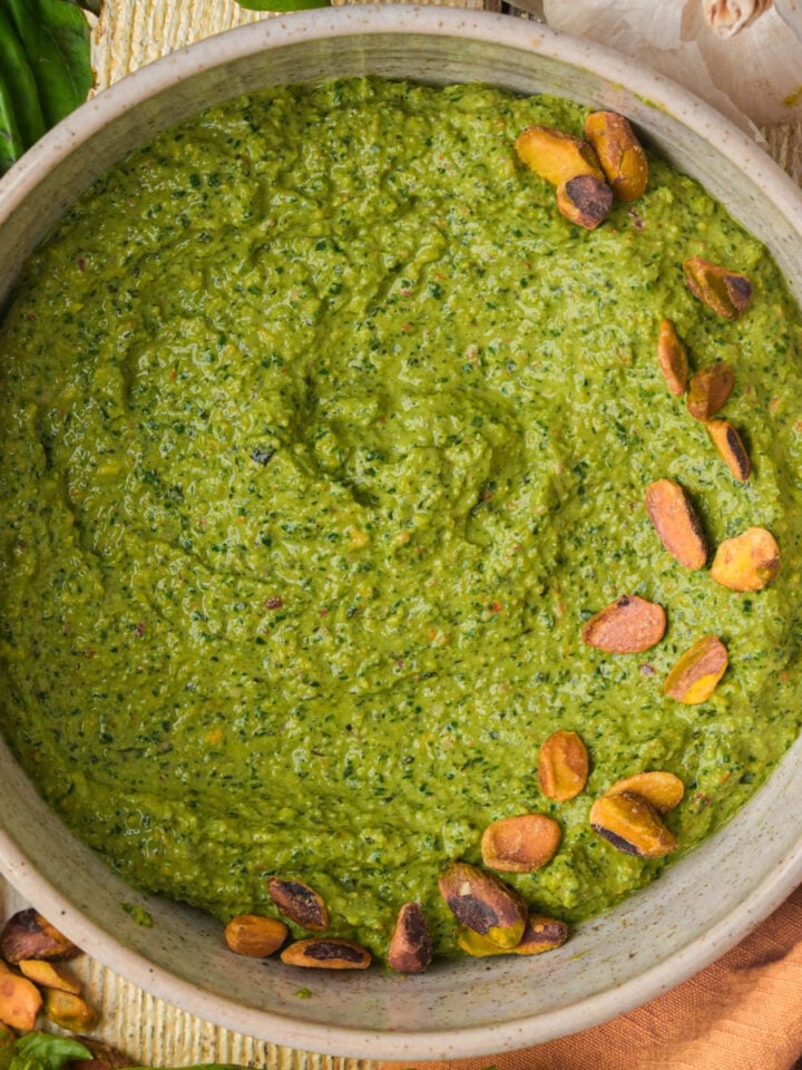 A bowl of pistachio pesto garnished with whole pistachios, surrounded by fresh basil leaves and a bulb of garlic on a wooden surface.