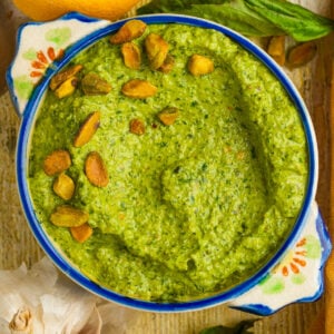 A bowl of pistachio pesto sauce garnished with pistachios, surrounded by fresh basil leaves, a lemon, and a garlic bulb on a rustic wooden surface.