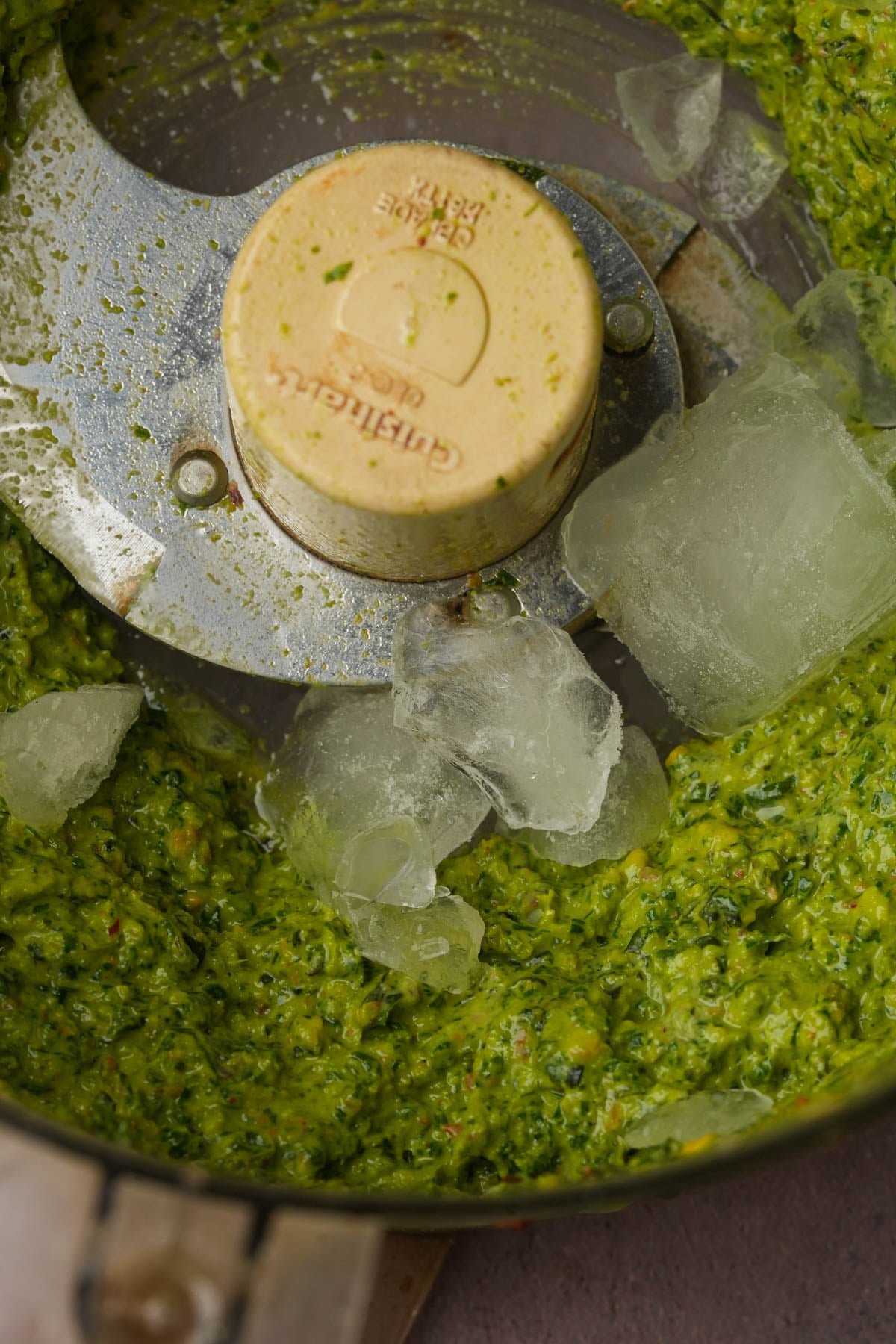 A food processor containing a bright green pistachio pesto with several ice cubes sitting on top of the pesto.