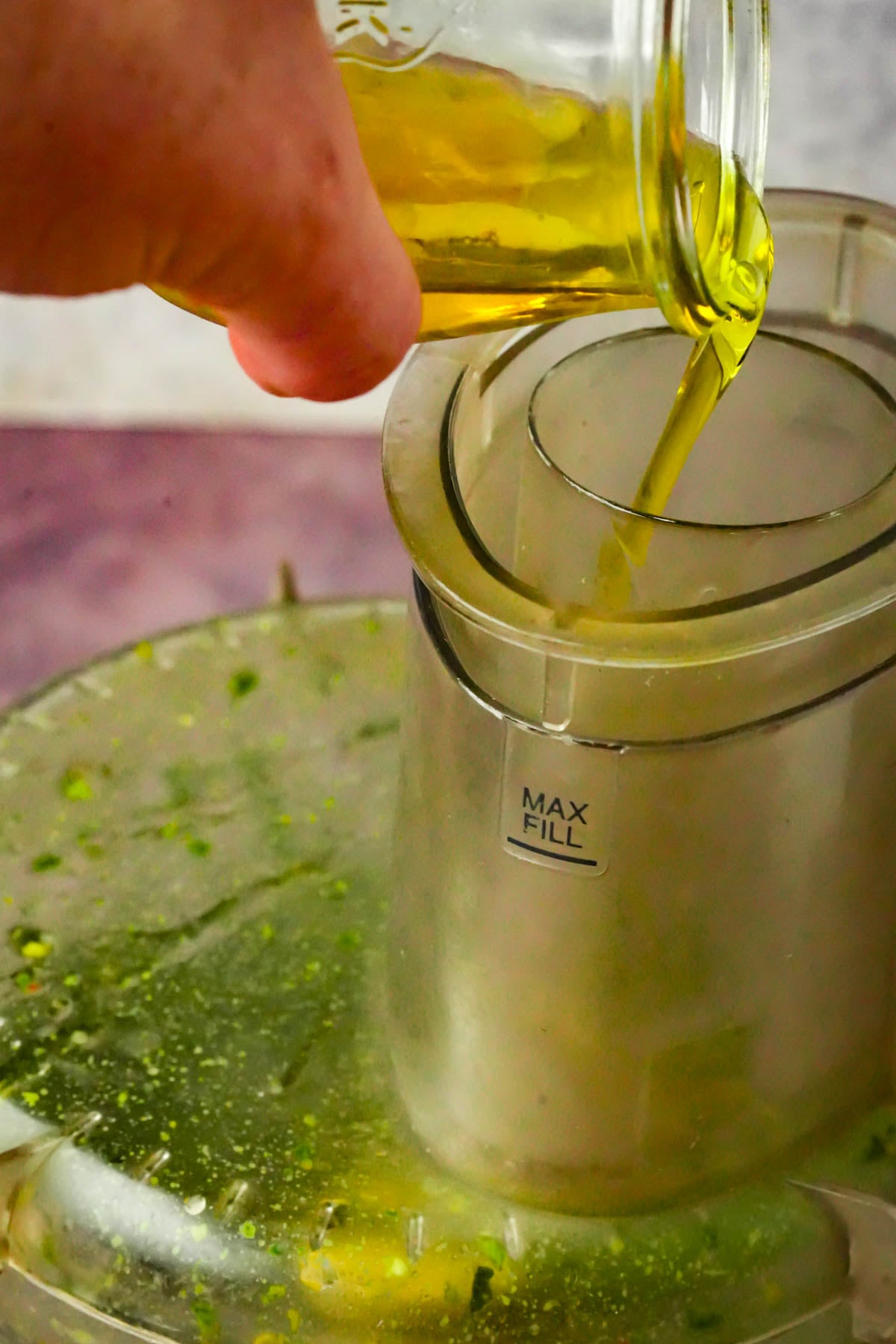 A hand pours olive oil from a glass jar into the feed tube of a food processor, which contains pistachio pesto ingredients and has a visible MAX FILL marking.