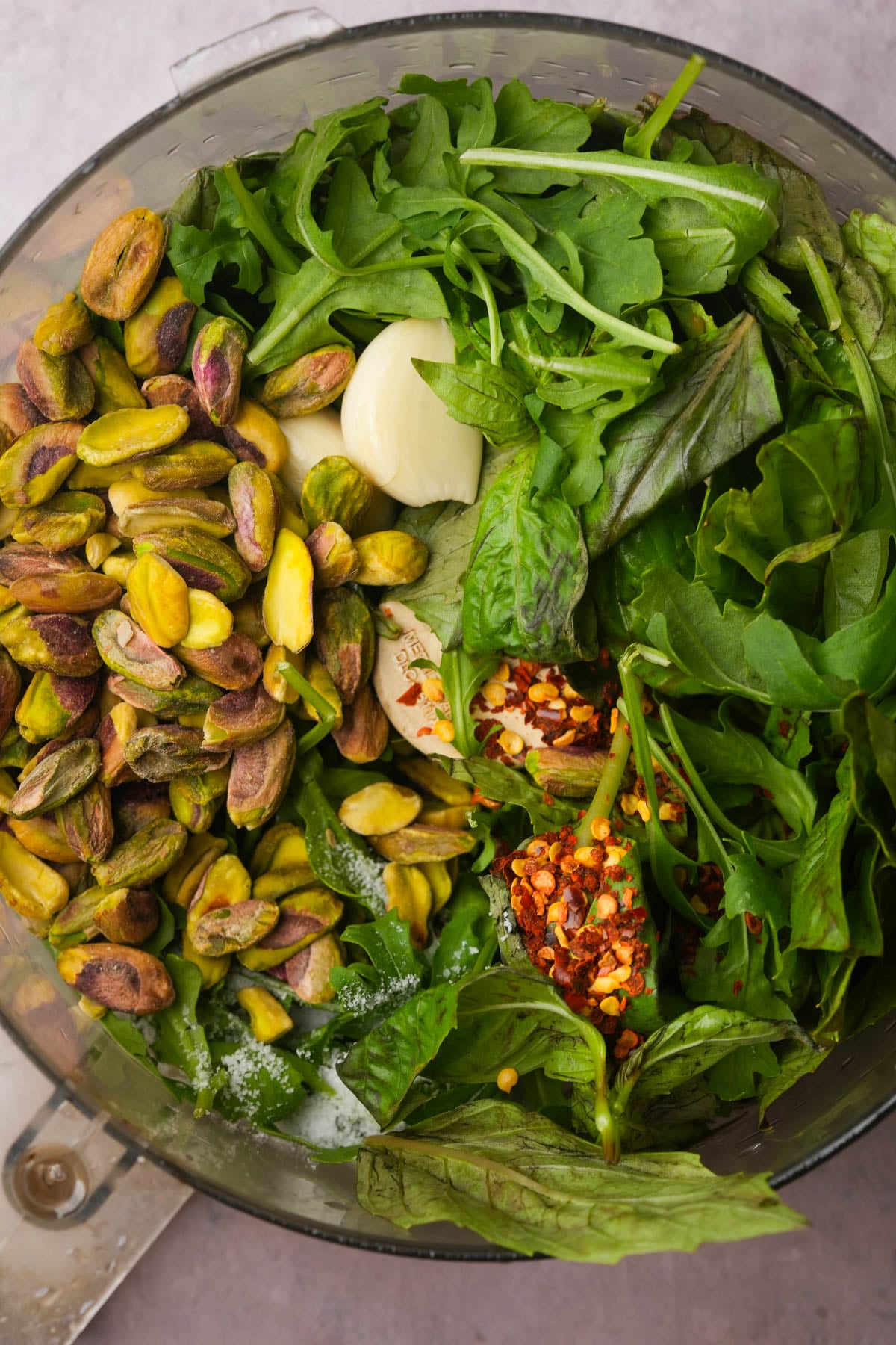 A food processor bowl filled with ingredients for a pistachio pesto recipe including fresh arugula, basil leaves, whole garlic cloves, shelled pistachios, red pepper flakes, and salt, ready to be blended into a pesto sauce.