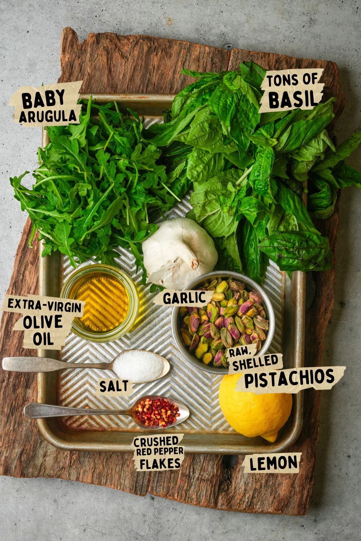 A tray on a wooden board holds labeled ingredients for a pistachio pesto recipe: baby arugula, lots of basil, a lemon, raw shelled pistachios, garlic, extra-virgin olive oil, salt, and crushed red pepper flakes.
