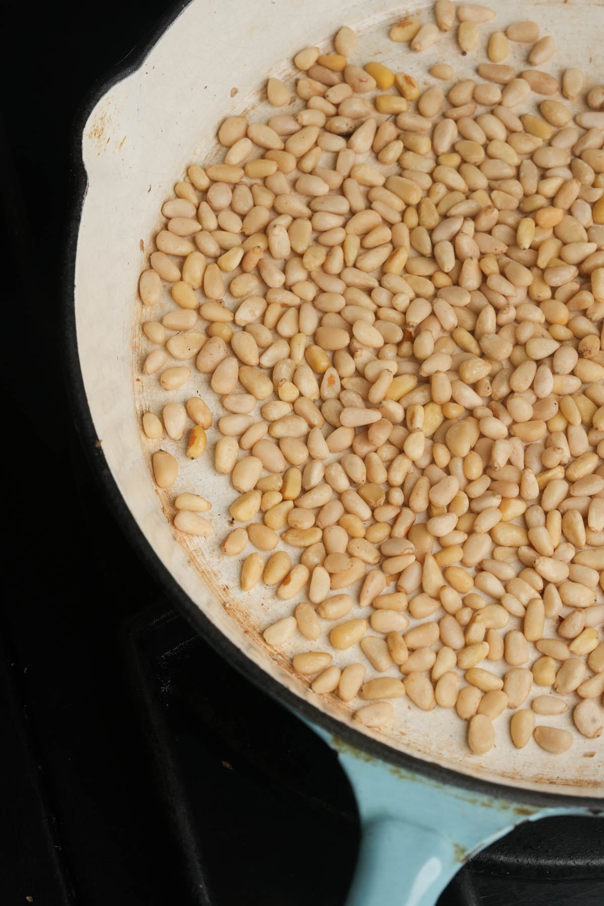 A close-up of pine nuts being toasted in a white skillet on a stovetop, with some nuts beginning to turn golden brown.