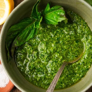 A bowl of spinach pesto garnished with fresh basil leaves, with a spoon in the bowl. Part of a lemon and a small dish of coarse salt are visible beside the bowl.